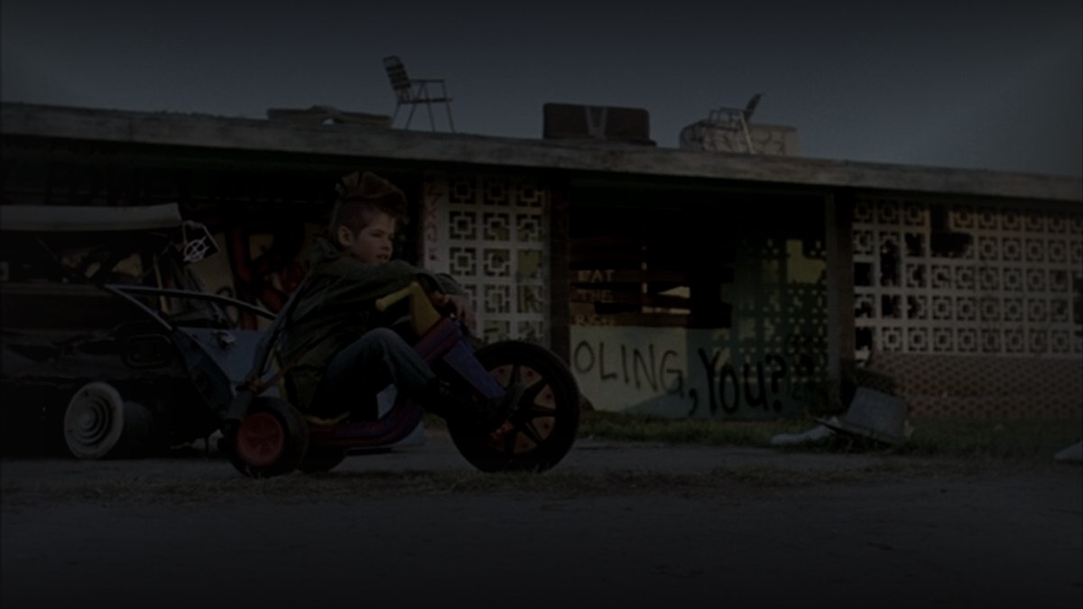 Still from Suburbia (1983) — letterboxd og:image still, cross-checked against three tmdb backdrop candidates; pre-darkened 0.75, baked through scripts/bake_dark_photo.py at factor 0.45, output luma 23.9, 1200x675
